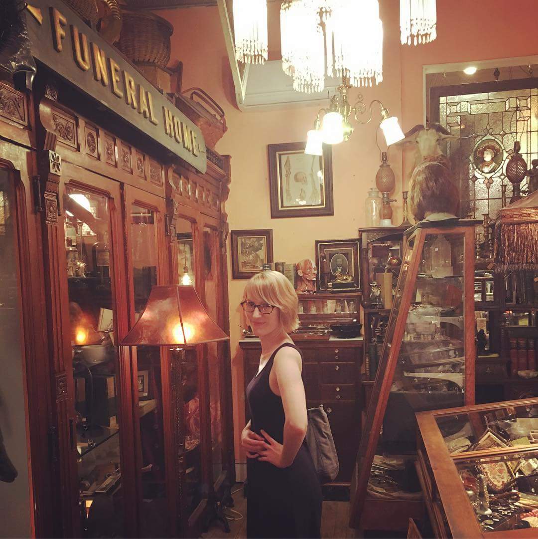 A woman stands next to a large wooden display case filled with antiques. 
